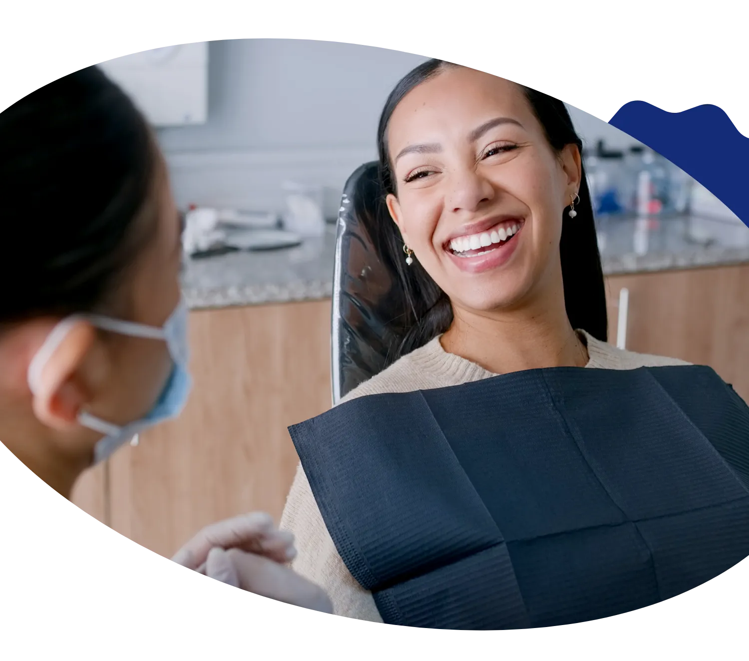 A patient smiles while sitting in a dentist's chair, talking to a dental professional wearing a mask, framed within an oval-shaped design with a decorative blue accent.